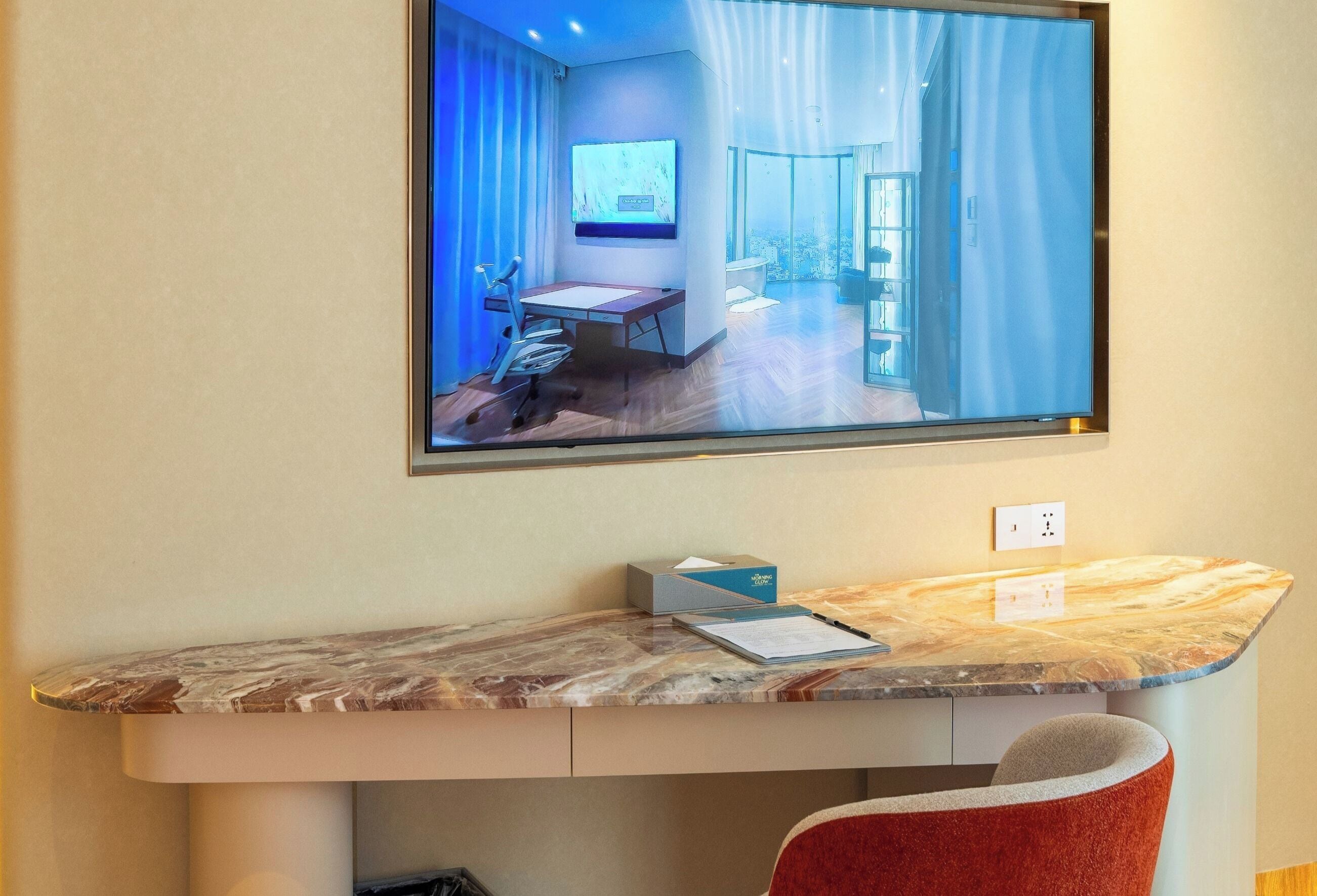 A hotel room with a large TV mounted on the wall, a desk with a chair, and a view of the city through the window.