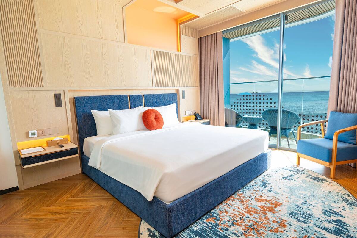Ein Hotelzimmer mit einem großen Bett, einem Schreibtisch mit Telefon, einem Sessel und Blick aufs Meer.