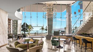 Un hall d’entrée spacieux avec un plafond incurvé, un lustre central, un coin salon avec des canapés et des fauteuils, et une vue sur la mer grâce à de grandes fenêtres.