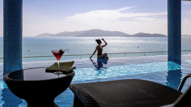 Une vue sur la piscine avec une personne dans l’eau, une boisson sur une table, et un panorama sur la mer et les montagnes.