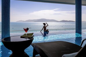 Vista de la piscina con una persona en el agua, una bebida en una mesa y vistas al mar y a las montañas.