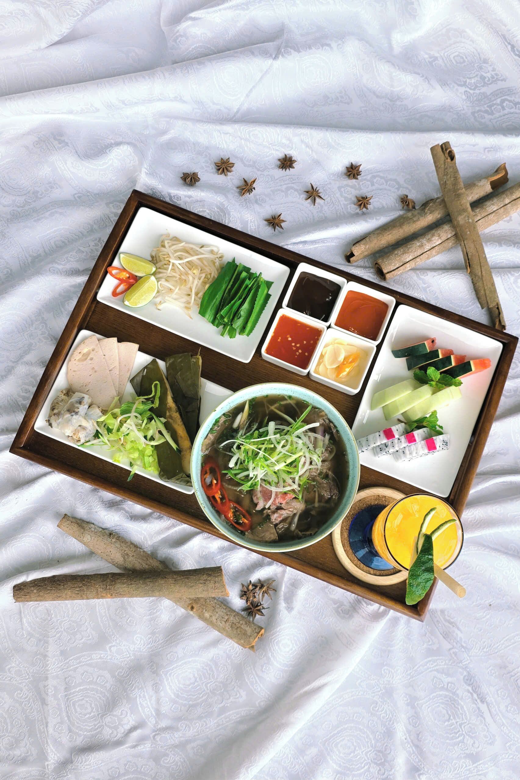 Un plateau contenant divers aliments et condiments, notamment des nouilles, des légumes et un bol de soupe, posé sur une nappe blanche à motifs.