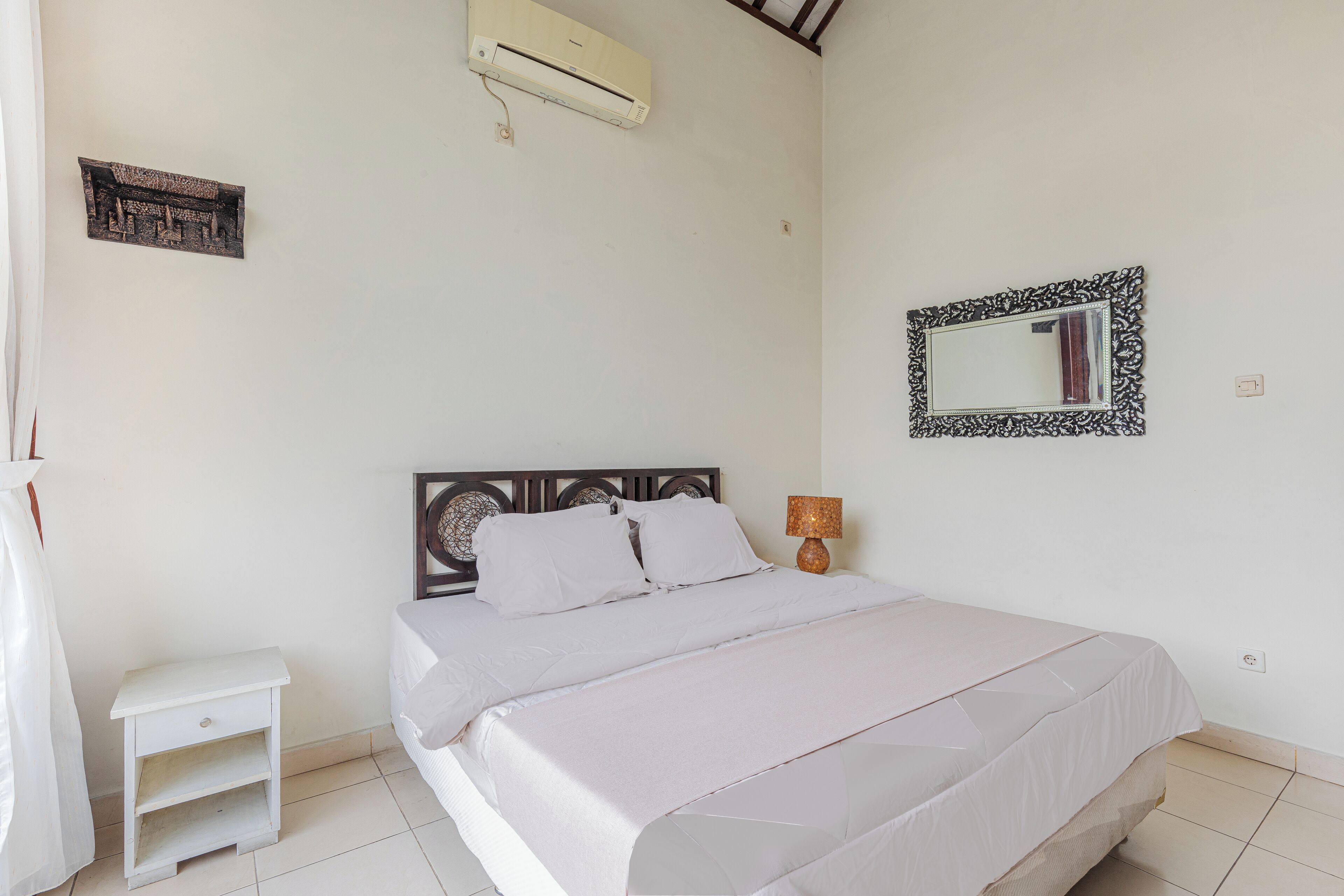 A bedroom with a bed, bedside table, lamp, air conditioner, and decorative mirror.