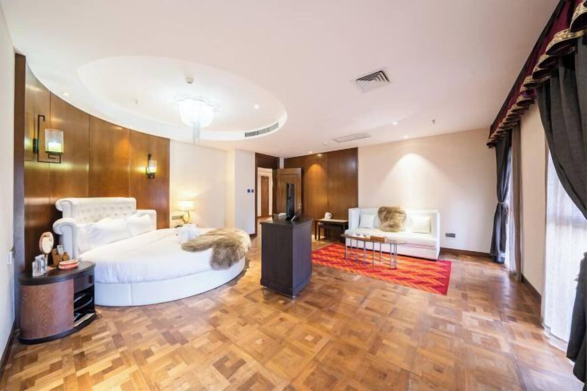 A hotel room with a curved bed, a sitting area with a sofa and armchair, a small table, and a large mirror.