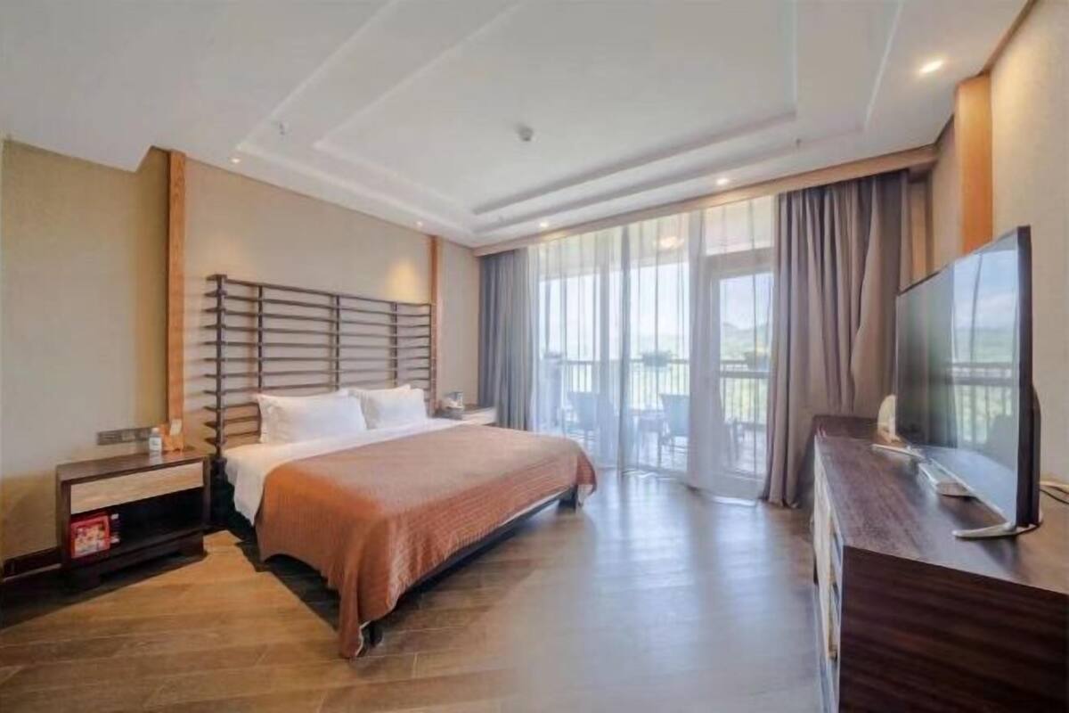 A hotel room with a large bed, a TV, a desk, and a balcony with curtains.