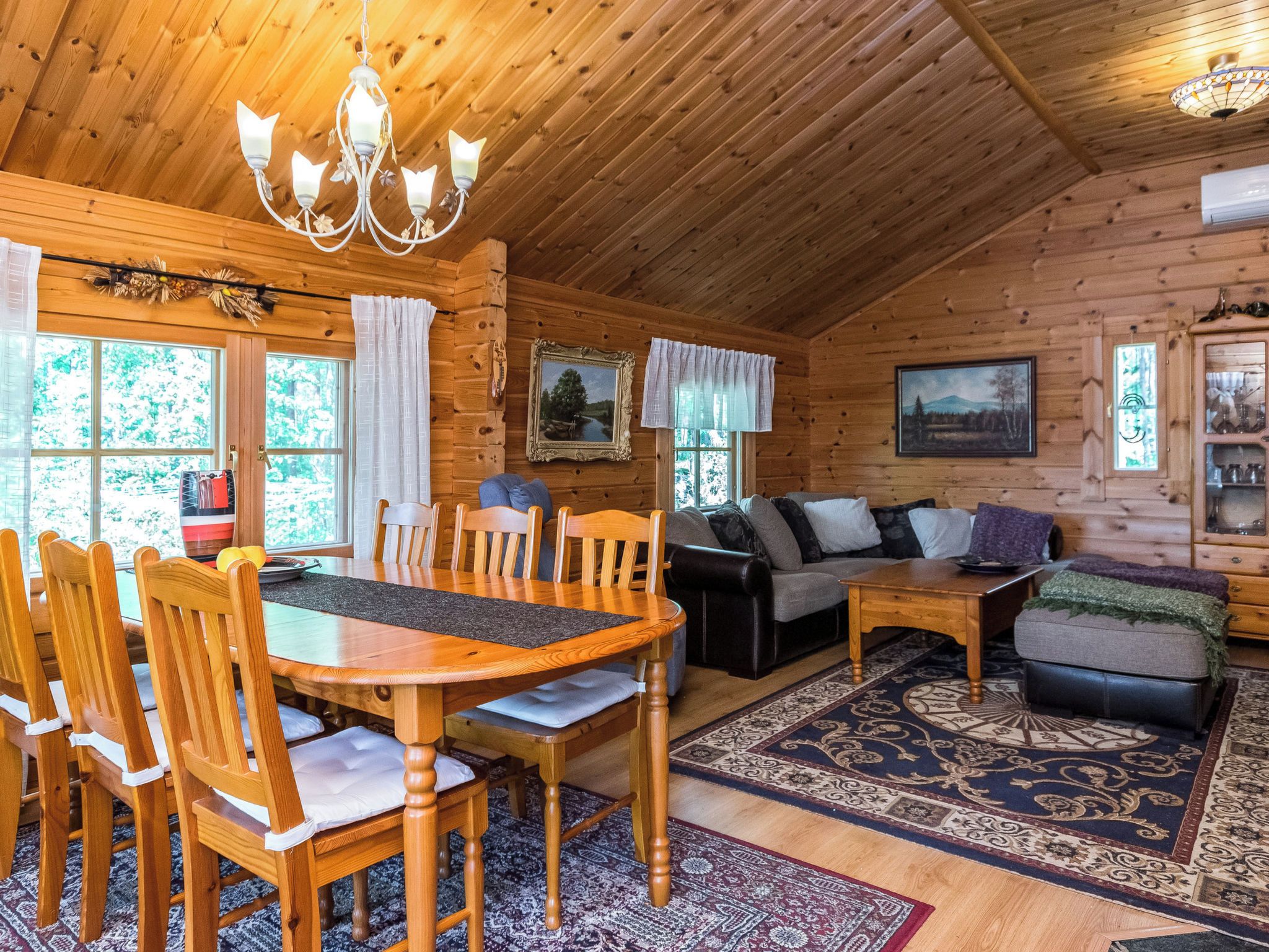 Ein Holzblockhaus-Interieur mit einem Esstisch, Stühlen, einer Couch und einem gemusterten Teppich.
