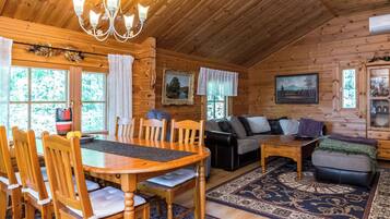 A wooden cabin interior with a dining table, chairs, a sofa, and a patterned rug.
