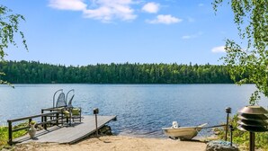 Eine Holzsteg, der sich in einen See erstreckt, mit einem Stuhl und Tisch darauf, einem an den Steg angebundenen Boot und einer bewaldeten Uferlinie im Hintergrund.