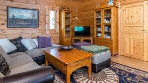 Ein gemütliches Blockhäuschen im Inneren, mit Holzvertäfelung, einer Couch, einem Couchtisch, einem Fernseher und Schränken mit Glastüren.