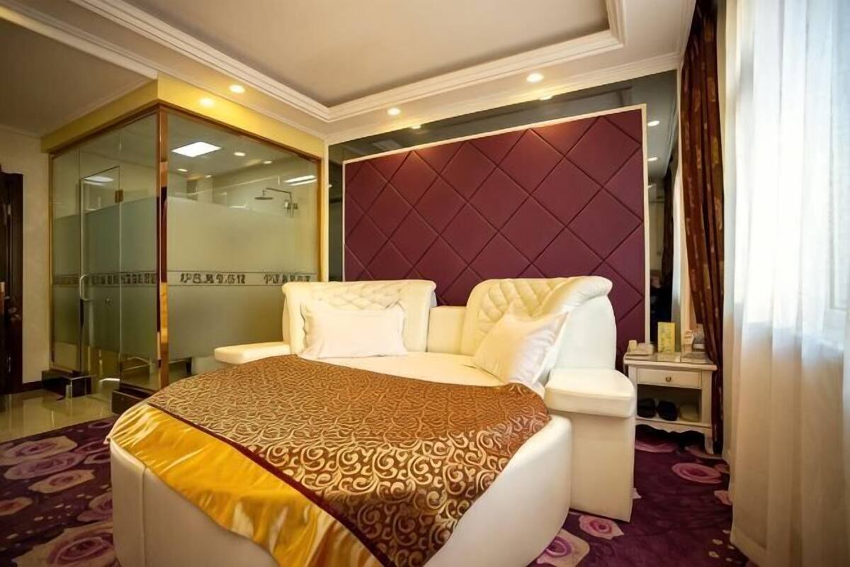 A hotel room with a bed, a bedside table, a mirror, and a glass shower enclosure.