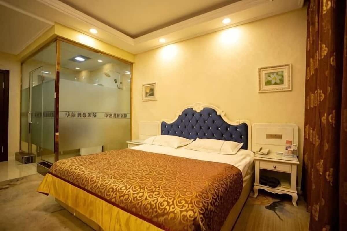 A hotel room with a large bed, a bedside table with a lamp, a glass door leading to a shower area, and a small table with a phone.