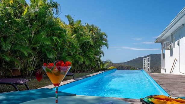 A poolside area with a clear view of the pool, surrounded by lush greenery and a white building. - Villa Malinina, swimming pool, sea view (Les Anses-d'Arlet.)