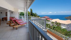 A balcony with sun loungers, a table, and chairs, overlooking a coastal view with houses and the sea. - Villa Malinina, swimming pool, sea view (Les Anses-d'Arlet.)