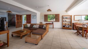 A spacious living room with a large sofa, a coffee table, and a dining area with a wooden table and chairs. There is a bookshelf and a television in the room. - Villa Malinina, swimming pool, sea view (Les Anses-d'Arlet.)