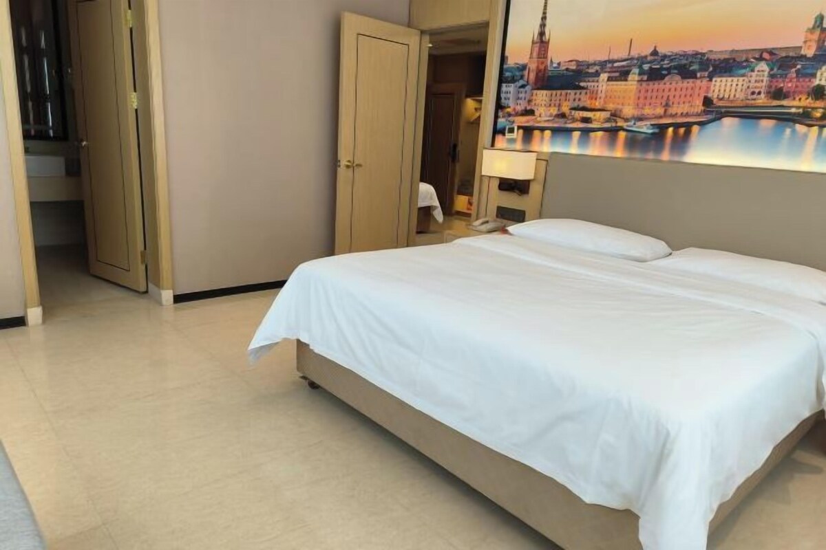 Quarto de hotel com cama, porta, pintura de uma paisagem urbana e mesa de cabeceira com abajur.