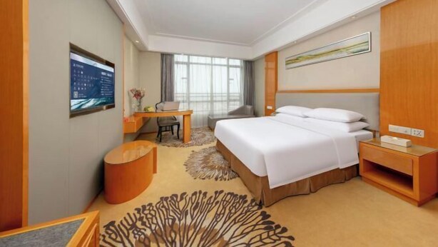 A hotel room with a bed, a desk, a chair, a small table, and a large window. - Vienna Hotel (Foshan Qiandeng Lake) (Foshan)