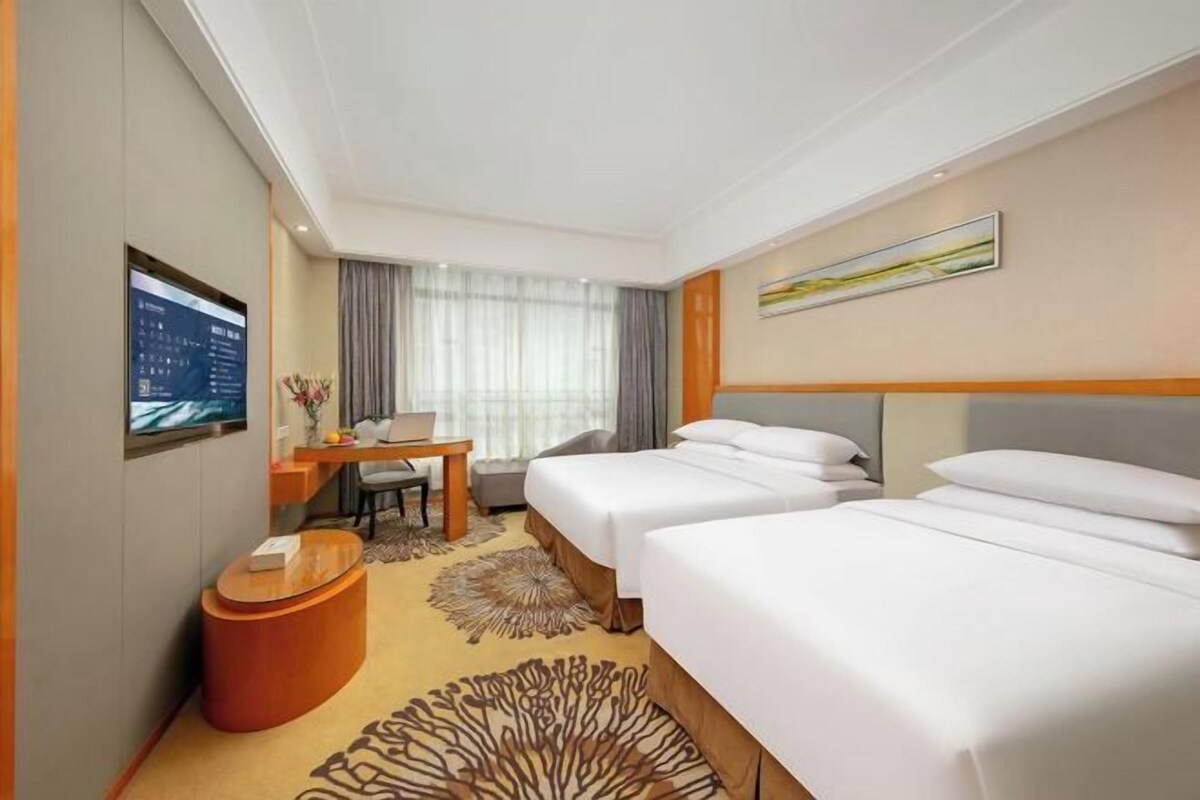 Une chambre d’hôtel avec deux lits, un bureau, une chaise, une petite table et une grande fenêtre avec des rideaux.