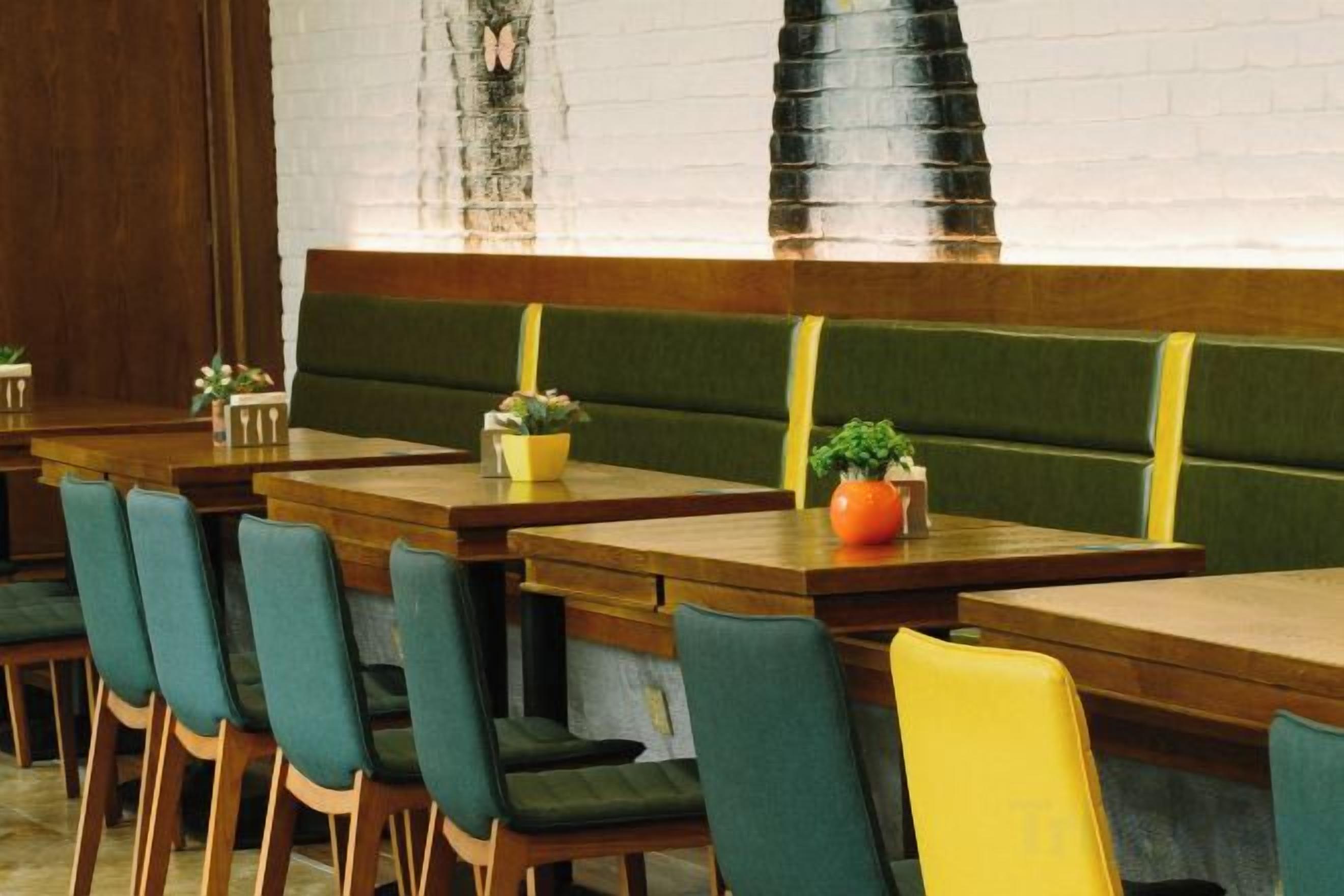 A restaurant interior with wooden tables, green and yellow upholstered booths, and potted plants.
