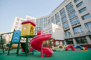 A playground with a red slide and climbing structures, set against a backdrop of a modern building with multiple windows. - Qixin Hotel (Hangzhou)