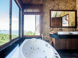 A modern bathroom with a large bathtub, a vanity with two sinks, a mirror, and a view of the sea.