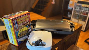 Una zona de cocina con una cocedora de arroz, una caja de fideos instantáneos y una regleta de enchufes sobre una mesa de madera.