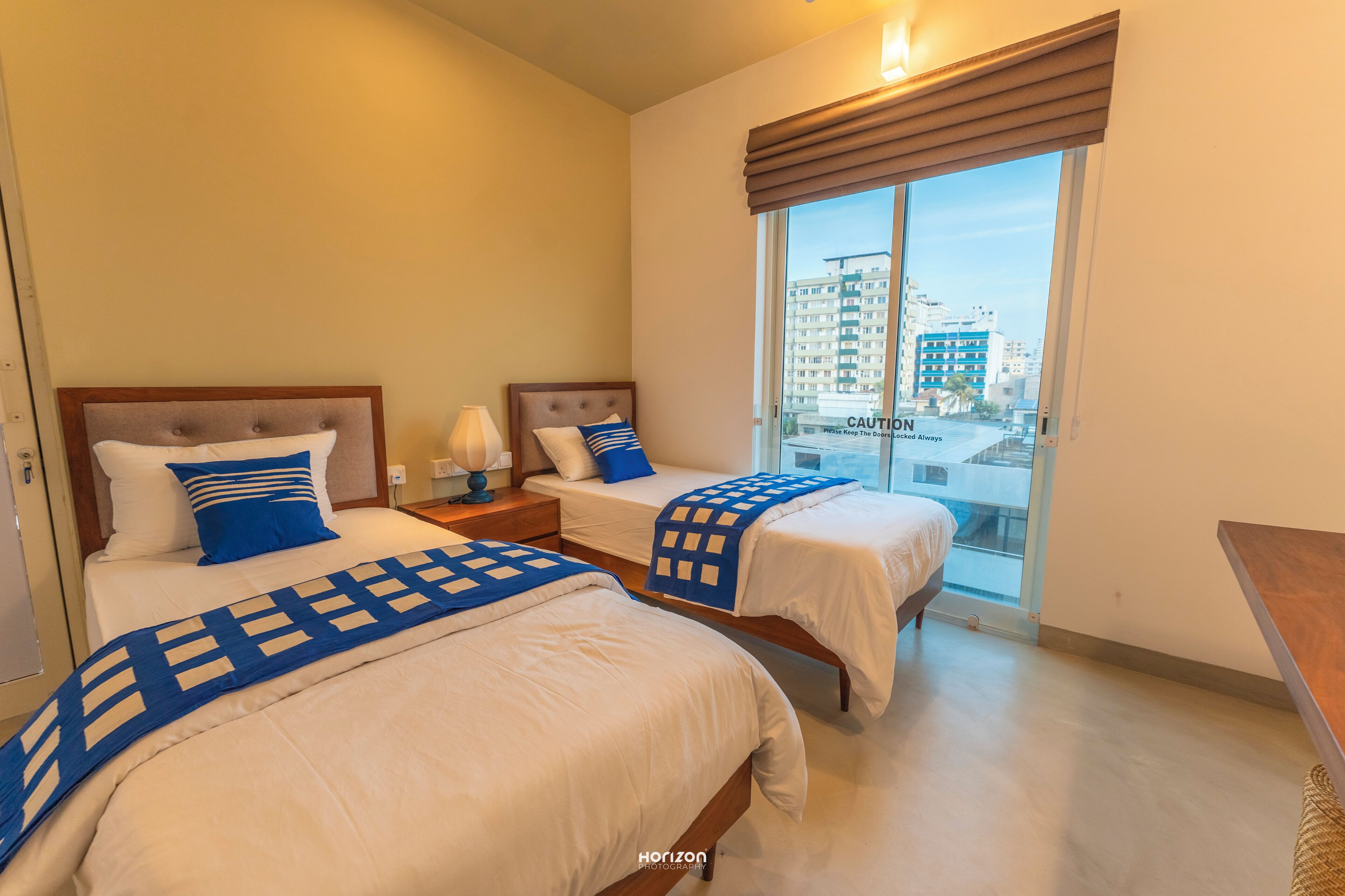 A hotel room with two beds, a nightstand, a lamp, and a view of the city through a large window.
