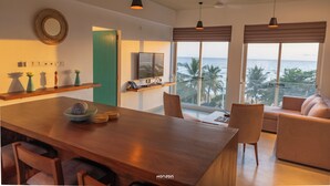 Una sala de estar moderna con una mesa de comedor de madera, asientos cómodos, ventanas amplias con vista al océano e iluminación colgante.