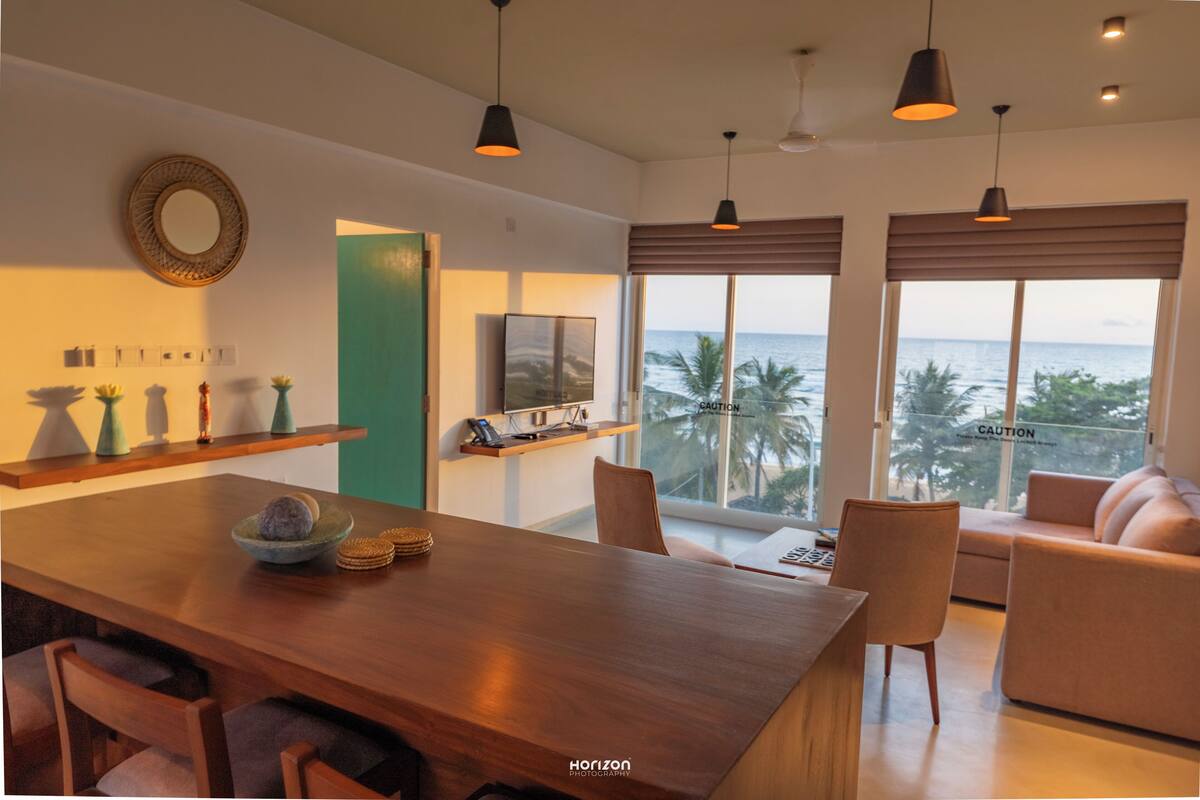 A modern living room with a wooden dining table, comfortable seating, large windows with a view of the ocean, and pendant lighting.