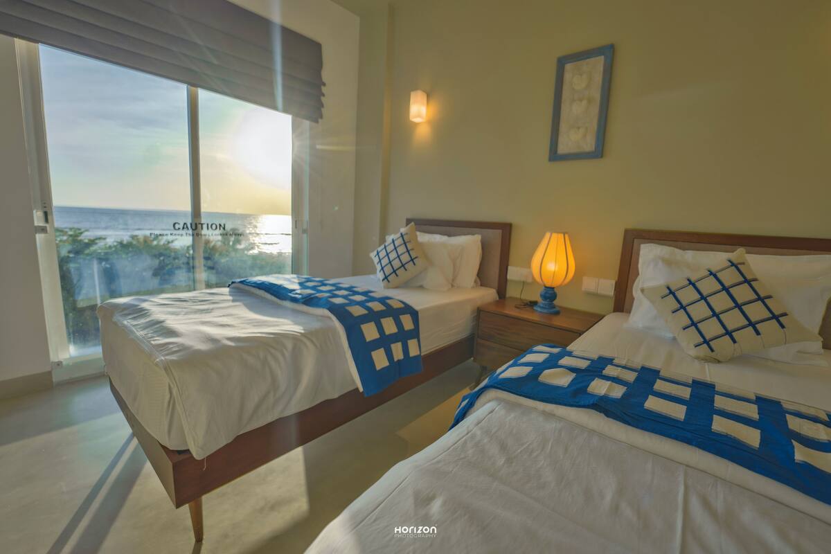 A hotel room with two beds, a nightstand, a lamp, a framed picture on the wall, and a view of the ocean through large windows.