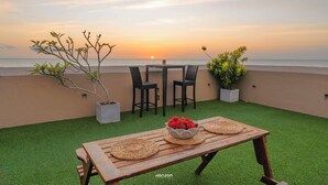 Una terraza con mesa de madera, dos sillas y vista al atardecer sobre el océano.