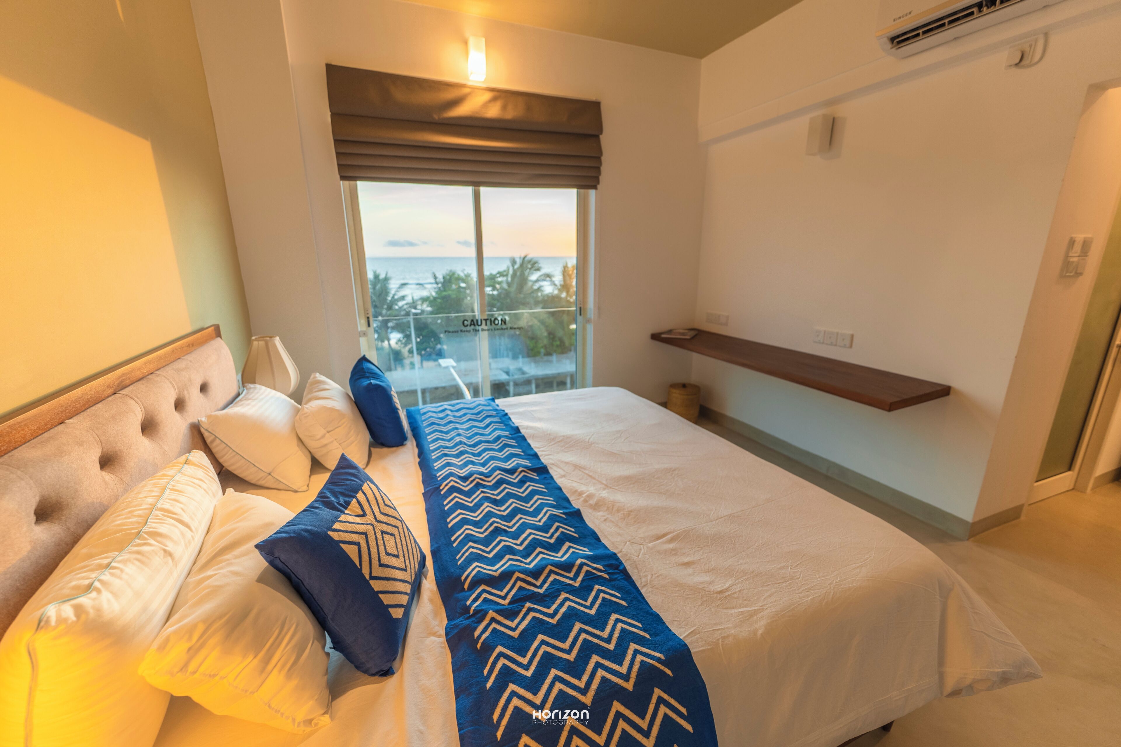 A bedroom with a bed, pillows, a bedside table, a window with a view, and a shelf.