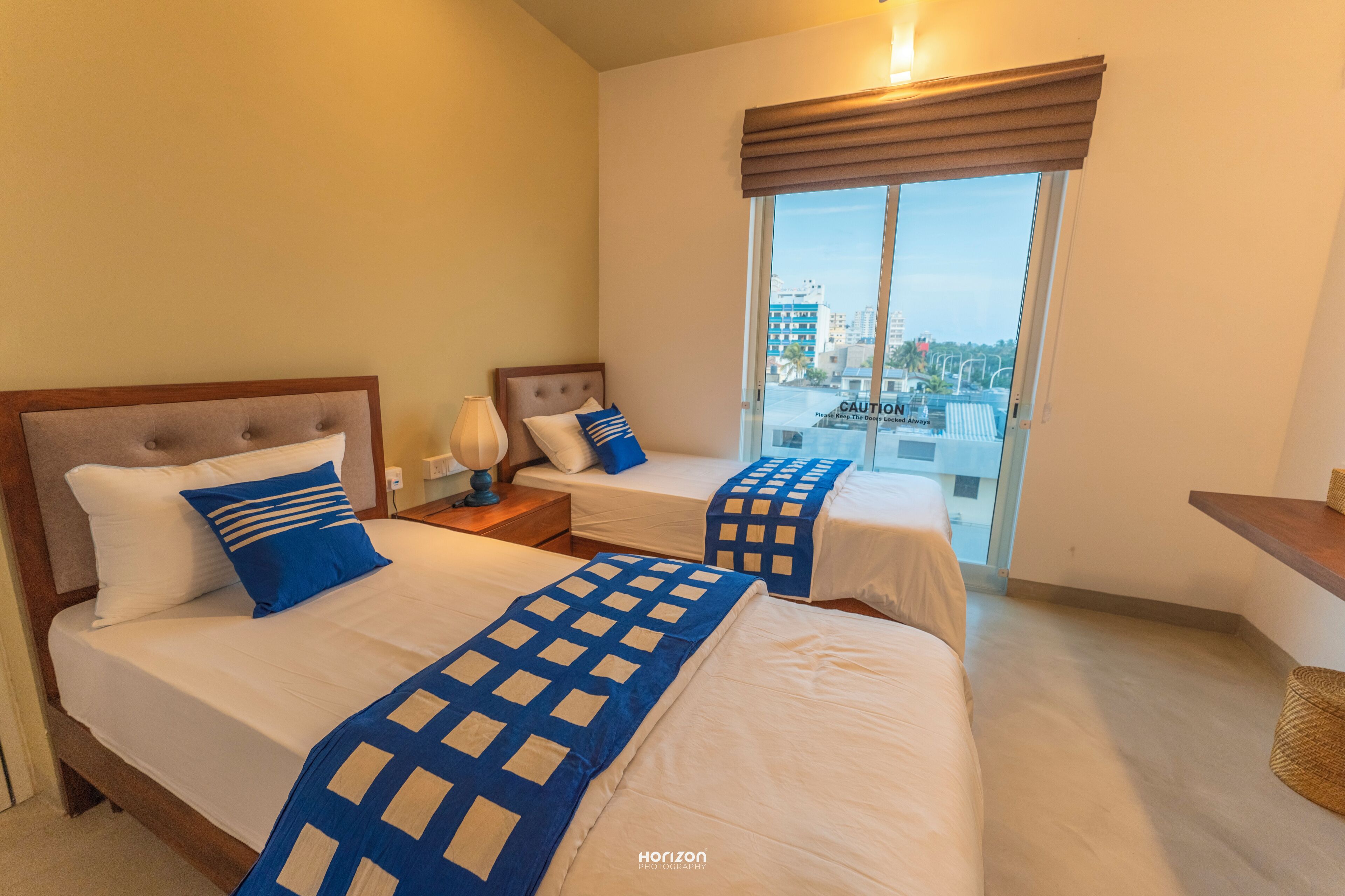 A hotel room with two beds, a nightstand, a lamp, a window with a view of buildings, and a wooden bench.