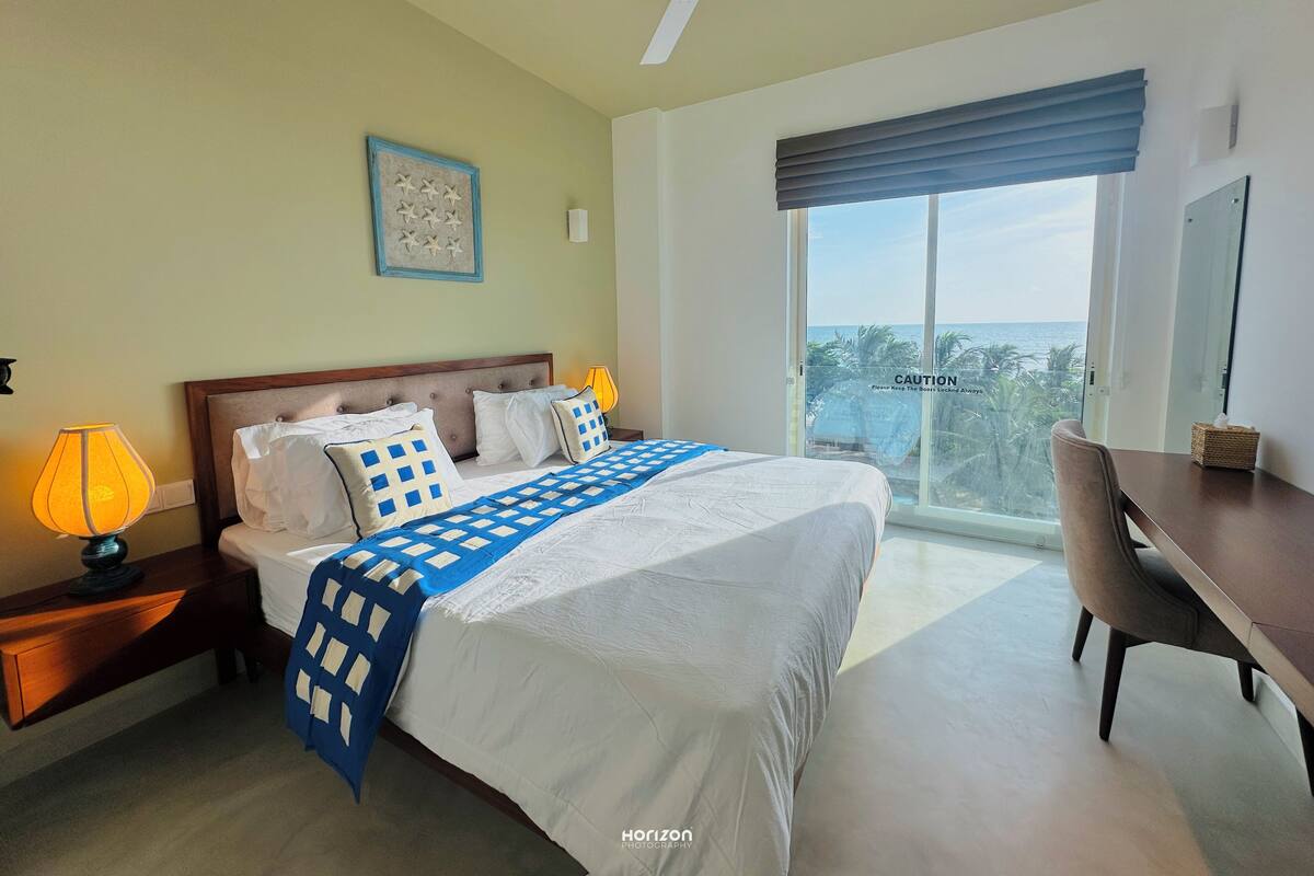 A hotel room with a large bed, a desk, a chair, a ceiling fan, and a view of the ocean through a large window.