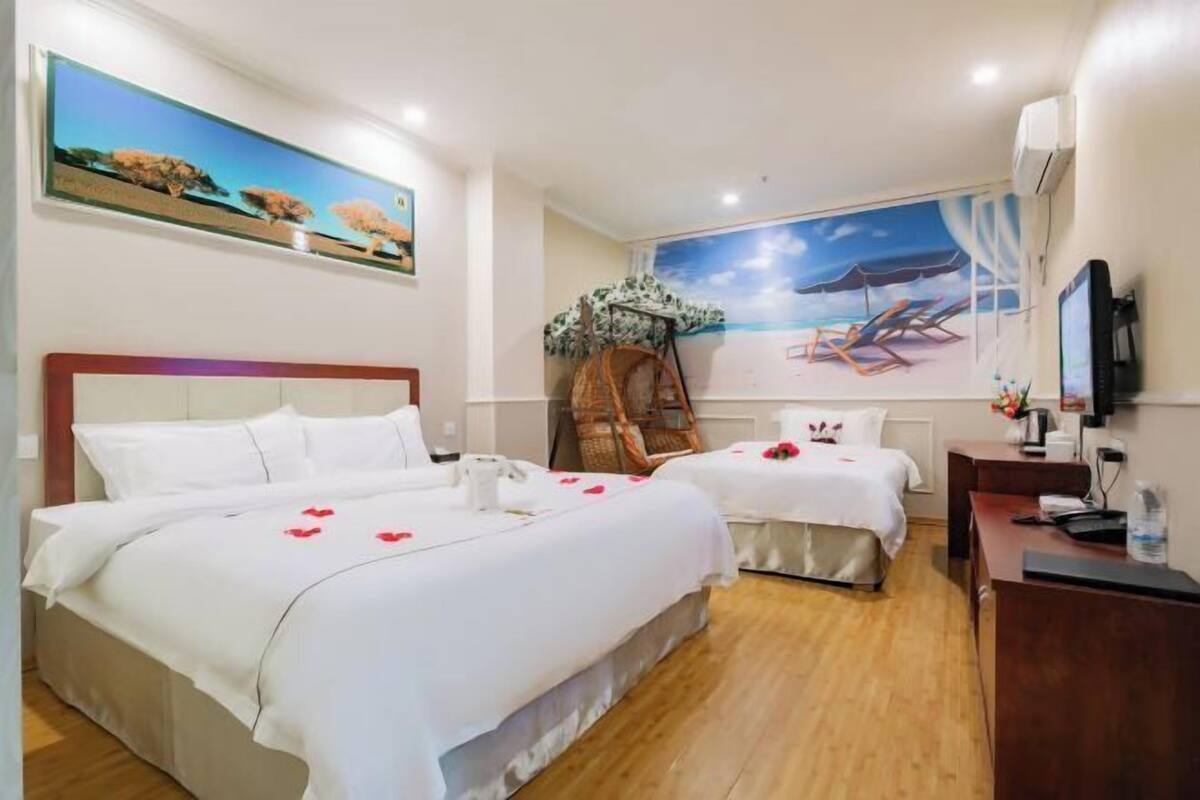 A hotel room with two beds, a painting of a beach scene, a wicker chair, and a desk with a television.
