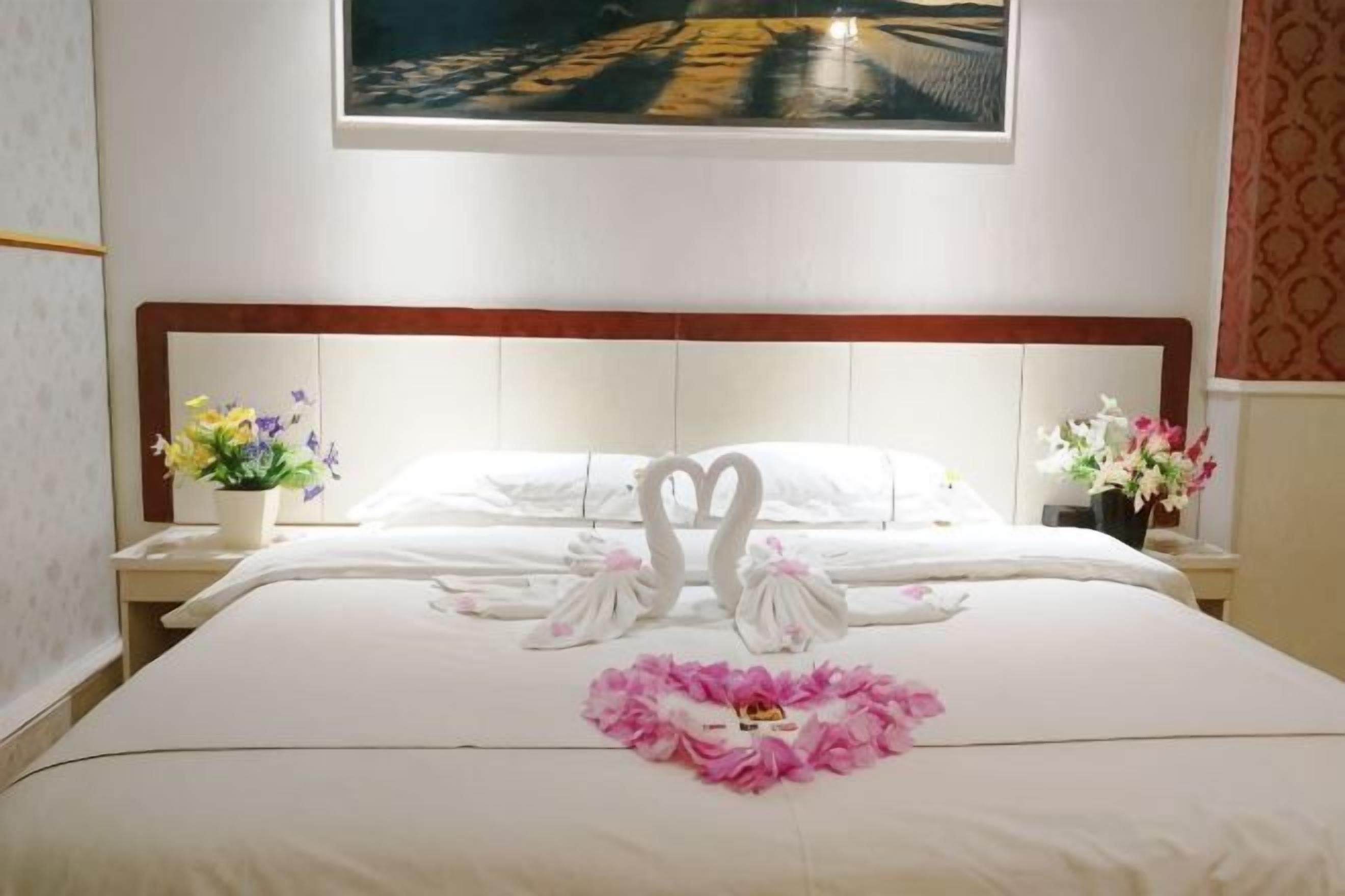 A hotel room with a neatly made bed, a painting above the bed, and two nightstands with floral arrangements.