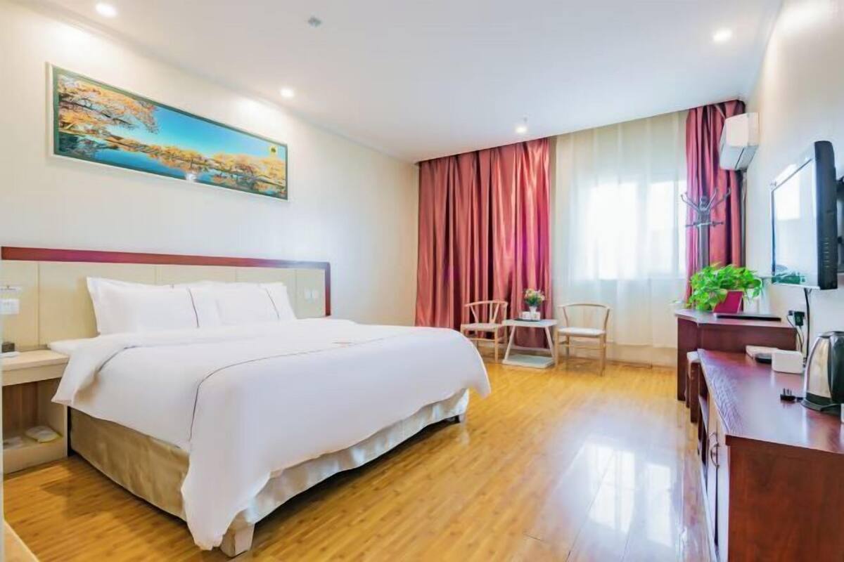 A hotel room with a bed, a desk with a chair, a small table with a plant, and a painting on the wall.