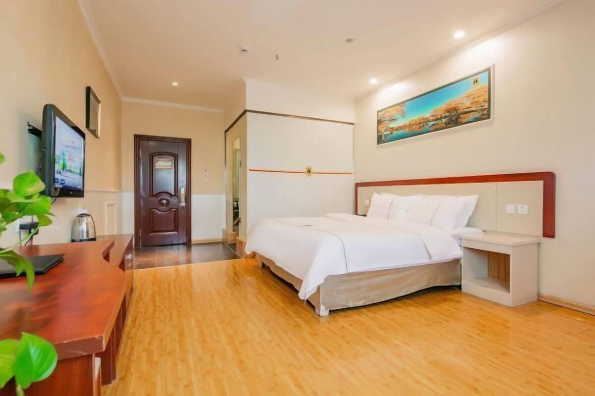 A hotel room with a bed, bedside table, TV, wooden floor, and a painting on the wall.