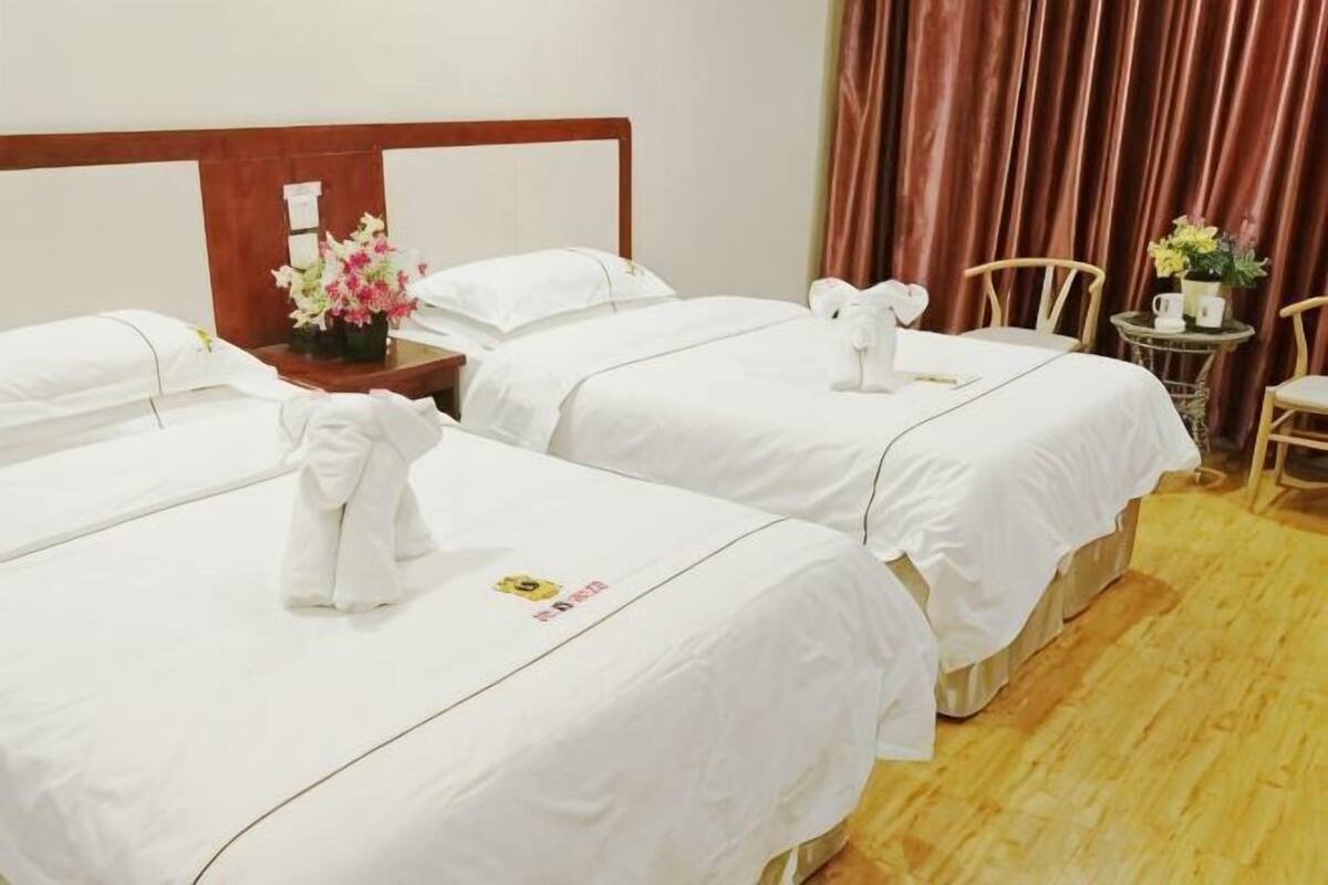 A hotel room with three beds, a wooden headboard, a small table with a vase of flowers, and a chair.