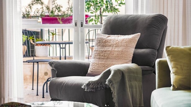 A modern living room with a grey sofa, a knitted throw pillow, and a view of a balcony with plants. - Ben Yehuda Green Charm by Sea N' Rent (Tel Aviv)