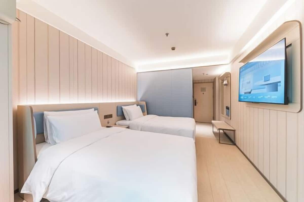 A hotel room with two beds, a TV mounted on the wall, and a view of a building through the window.