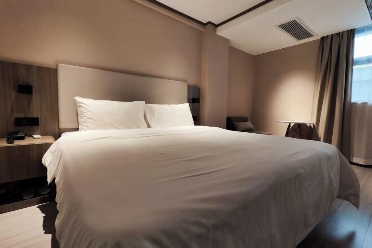 A modern hotel room with a large bed, a bedside table with a lamp, and a view of the outdoors through the window.
