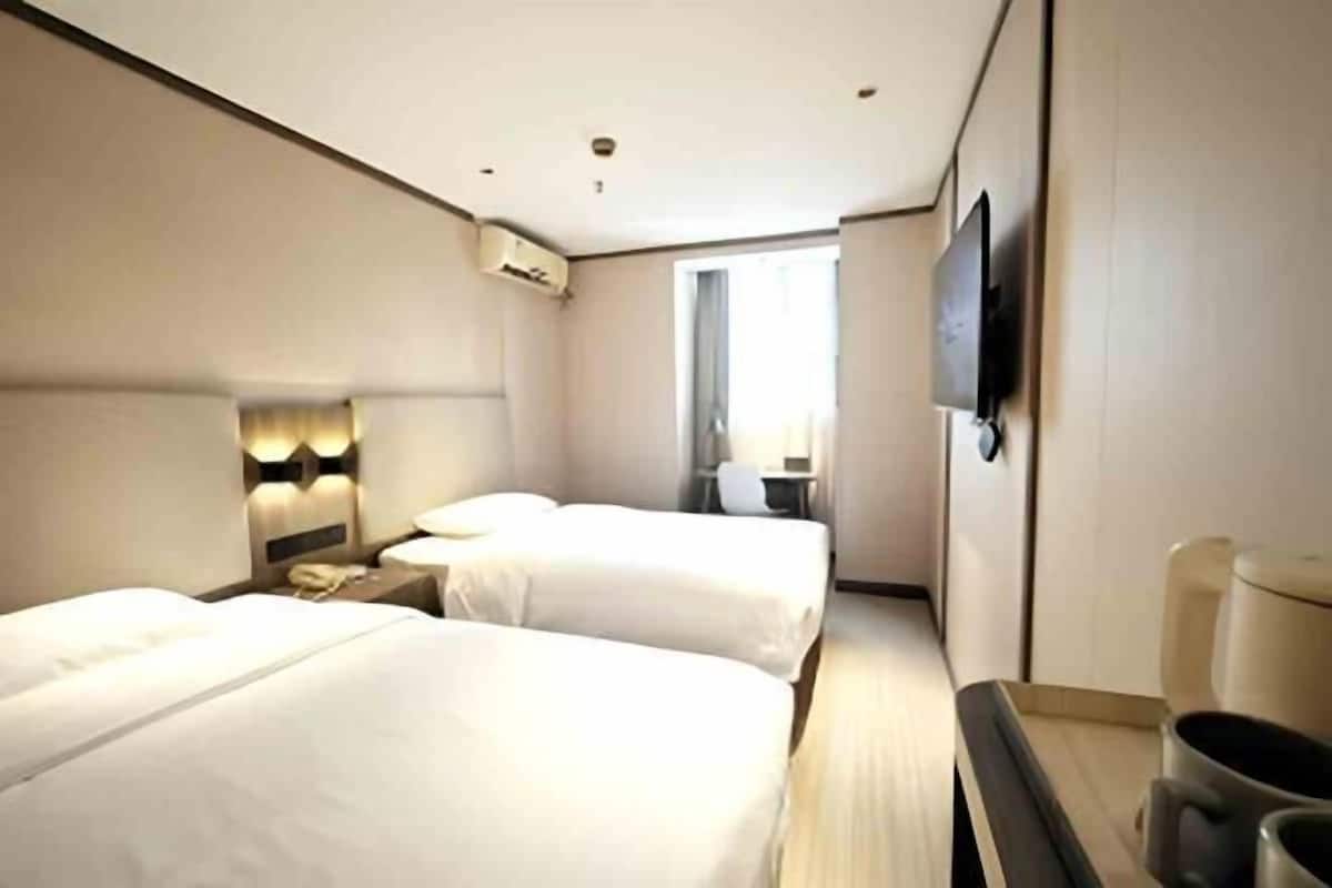 A hotel room with two beds, a TV mounted on the wall, a bedside table with a lamp, and a window providing natural light.