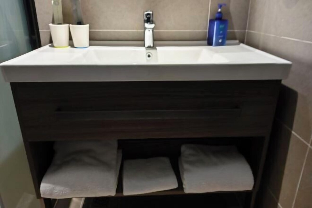 A bathroom with a white sink, a towel rack with three towels, a soap dispenser, and a toilet paper holder.