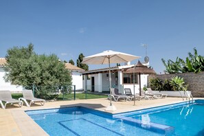 Une piscine entourée de chaises longues et de parasols, elle-même bordée d’un jardin clôturé et d’un bâtiment doté d’un toit en tuiles.