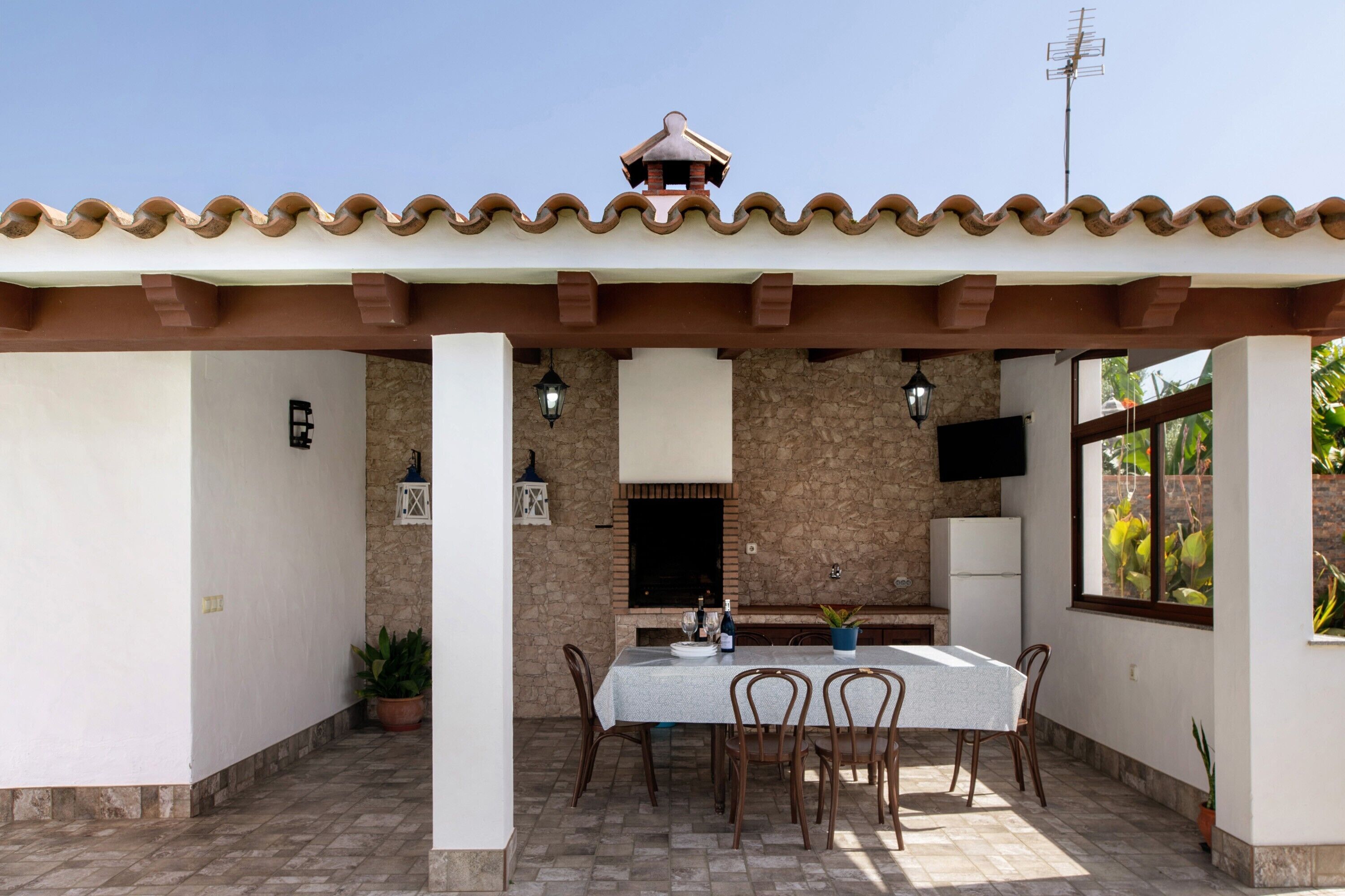 An outdoor patio with a table and chairs, a stone wall, a TV mounted on the wall, and a tiled roof.