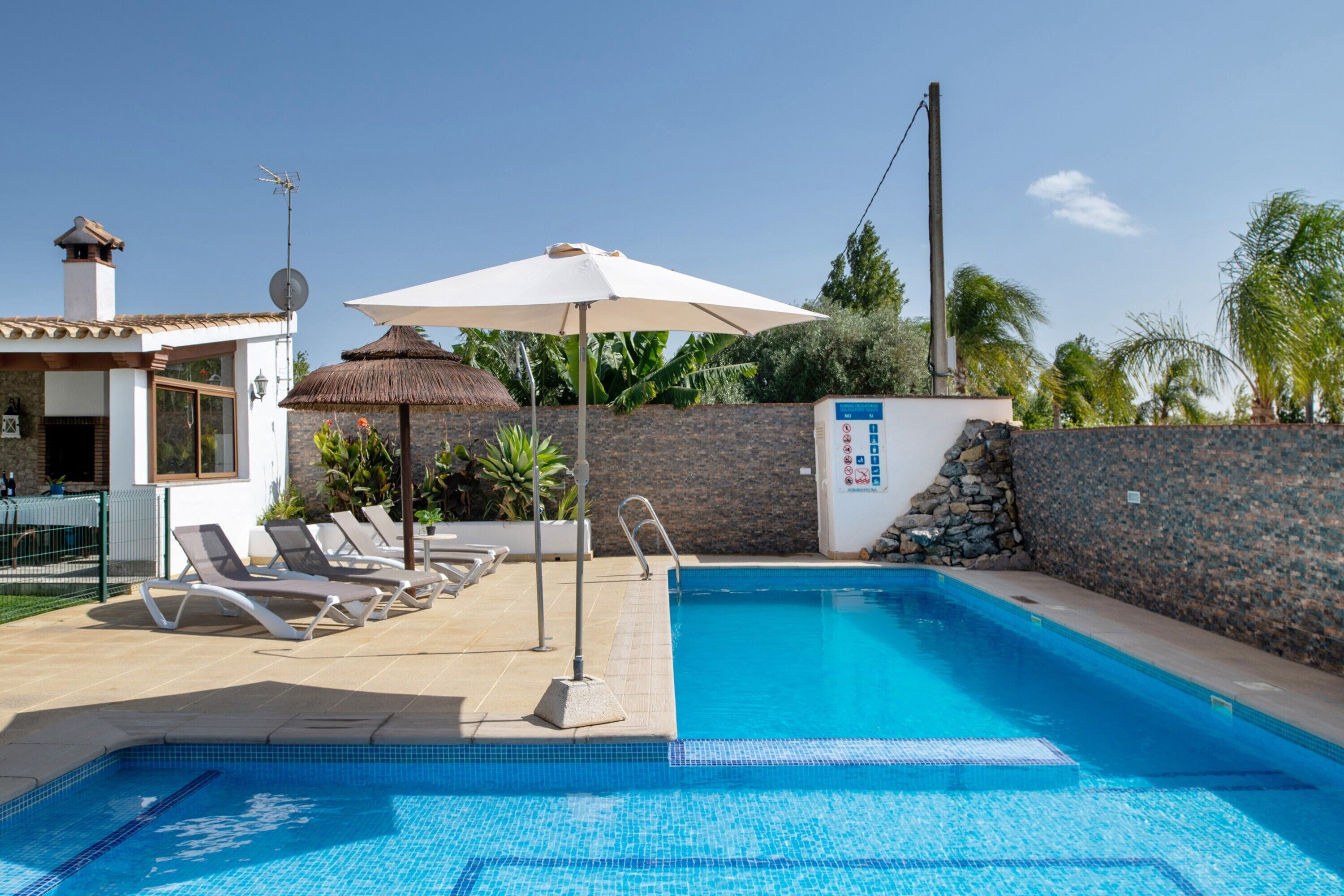 A swimming pool with lounge chairs and a sun umbrella, surrounded by a stone wall and vegetation.