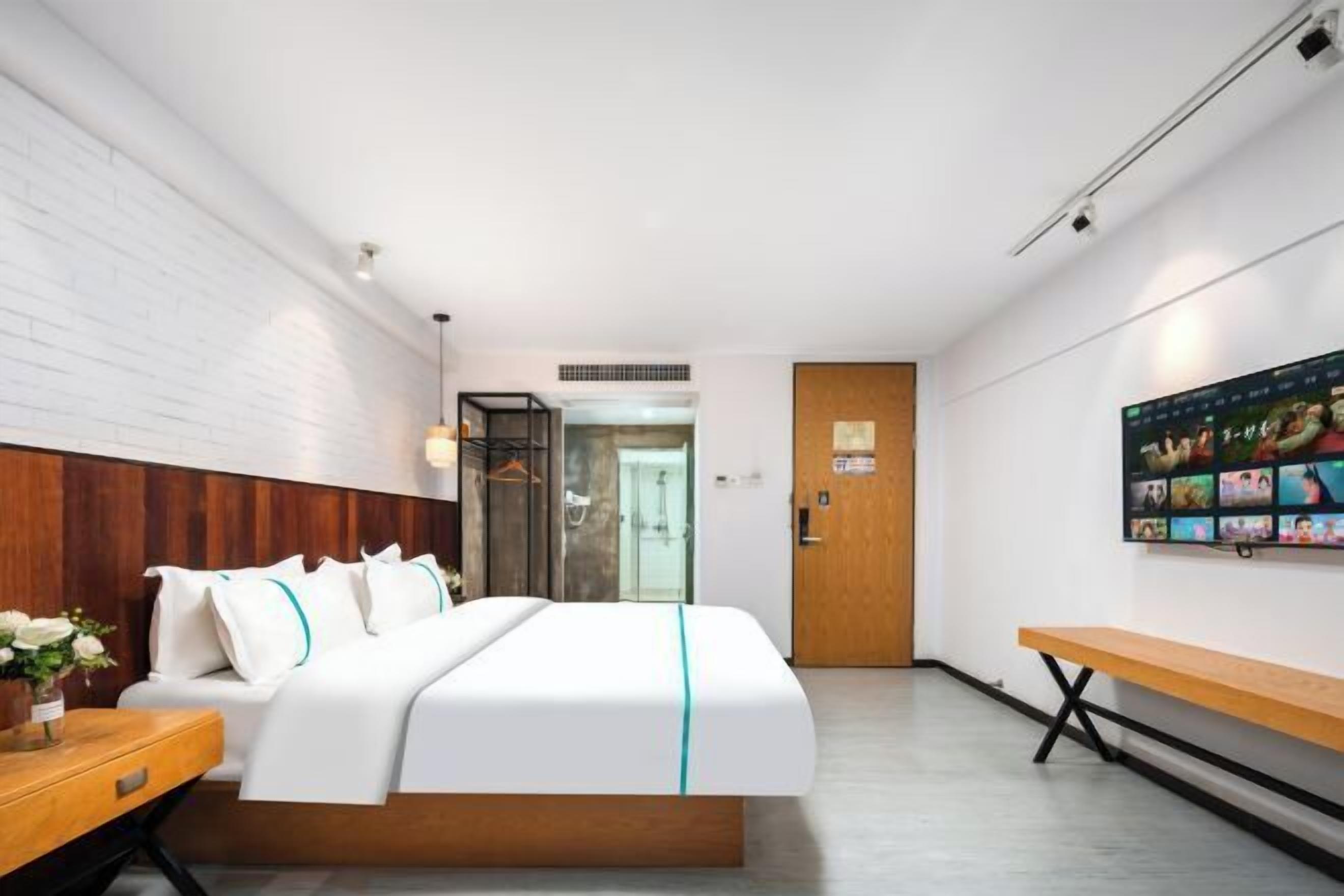 A modern hotel room with a bed, wooden headboard, a bench, a wall-mounted TV, and a door leading to a bathroom.