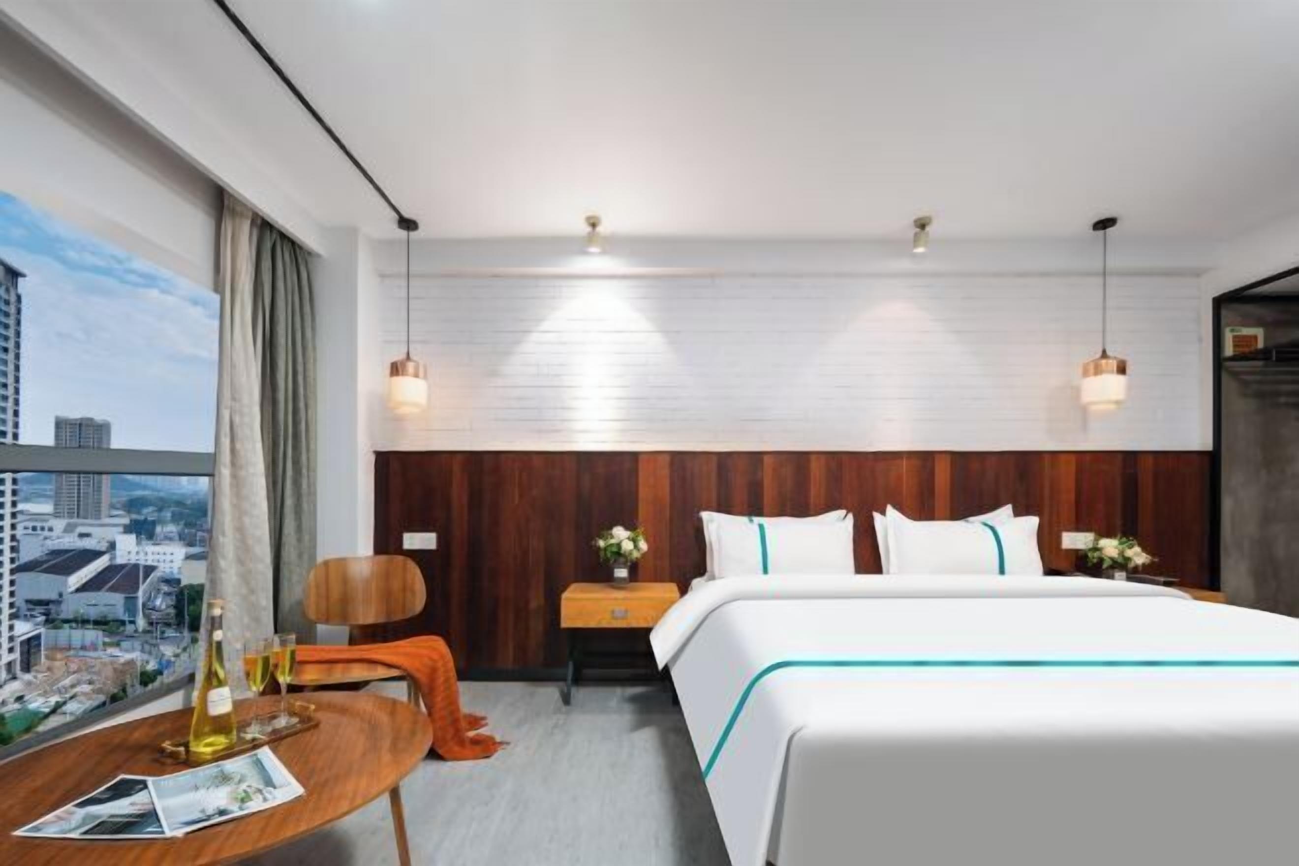 A hotel room with a bed, a wooden nightstand, a table with a bottle and glasses, a chair, and a city view.
