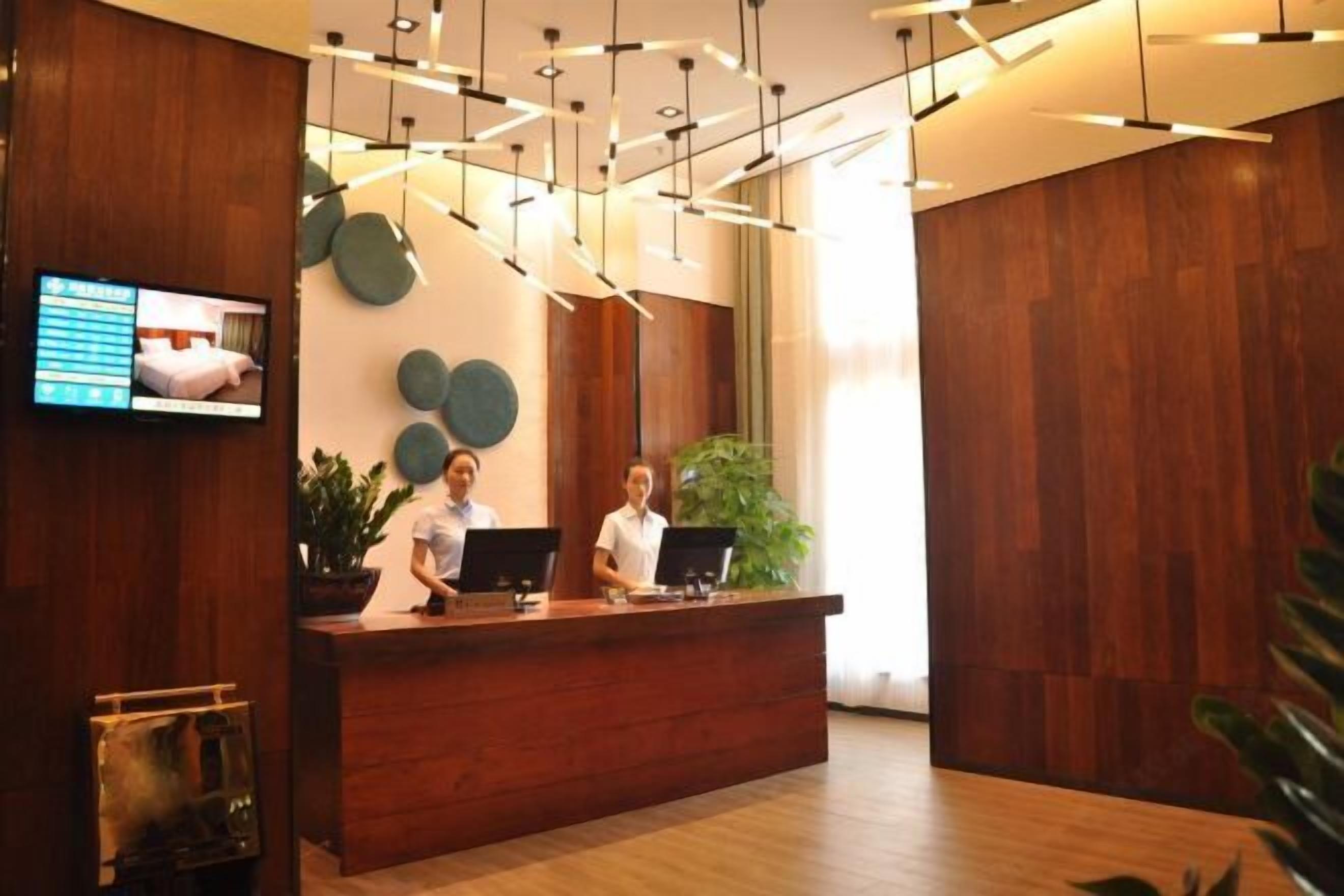 Una recepción con dos empleados, una pantalla digital que muestra opciones de habitaciones y un interior moderno con paneles de madera y luces colgantes.