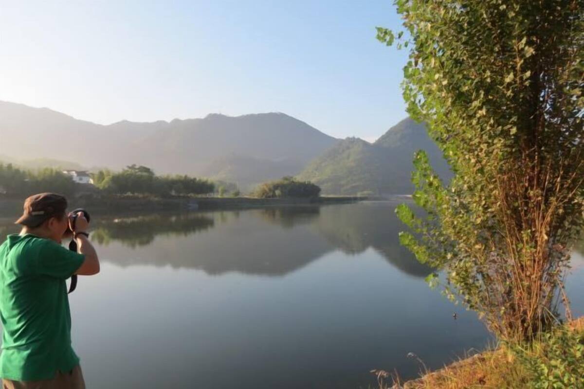 A person taking a photo by a calm lake with mountains in the background.